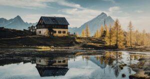 The 12 Best US Regions to Be In If Society Collapses A rustic cabin and mountains reflected in a serene alpine lake under a clear sky.