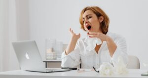 Why Millions of Americans With ME/CFS Still Don't Have a Diagnosis A woman in white yawning while working at a desk, indicating fatigue.