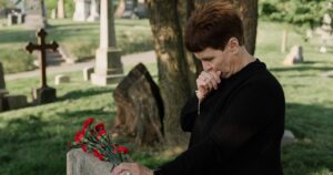 10 Mistakes to Avoid After Your Partner Passes Away (for Those Over 60) Senior woman in black at a cemetery, grieving by a gravestone with red flowers.