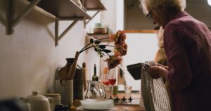 Elderly woman engaged in dish drying in a cozy, homey kitchen setting with shelves and flowers.