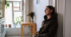 12 Signs of Emotional Neglect in Adulthood and How It Shapes Behavior Pensive woman in a hoodie sitting indoors by a window, capturing a moment of reflection.