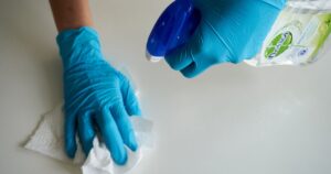 14 Things You Should Never Use Disinfecting Wipes On at Home Close-up of hands wearing gloves cleaning a surface with disinfectant spray.