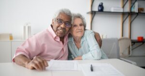 6 Smart Ways to Boost Your Social Security Payments in 2026 Elderly couple smiling and hugging while looking at documents on table indoors.