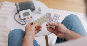 Close-up of elderly man sitting sofa at living room measures blood pressure with device. Senior male holding pills and drops drugs. Health and self-care concept, copy space