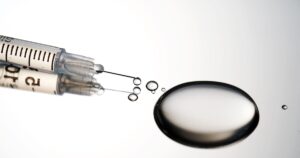 Injection syringe with a drop of drug on a stainless steel background.