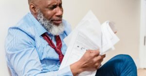 African American man in distress with paperwork, sitting against a wall indoors.