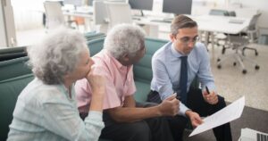 Some Couples Collect Over $100,000 a Year From Social Security — Here's How Business professional consults elderly clients in an office setting. Collaborative discussion, paperwork visible.