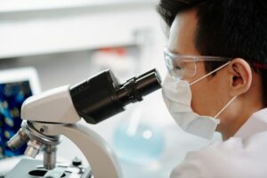 Bill Gates Says a Man-Made Virus Could Be Our Next Big Crisis Scientist wearing face mask and goggles examining samples through a microscope in a lab.