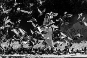Black and white photo of a woman walking through a flock of flying pigeons outdoors.