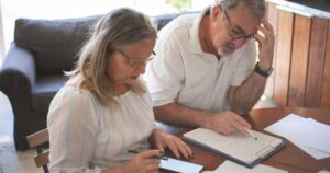 Why Owning a Paid-Off Home May Not Protect You in Retirement Elderly couple reviewing bills and documents at home, focusing on finances and technology.