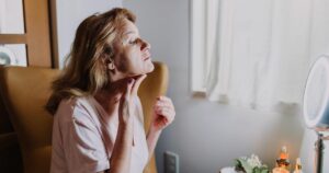 A mature woman examining her skin in a well-lit room with skincare products nearby.