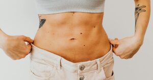 A woman shows weight loss progress by holding loose pants around her waist, highlighting tattoos.