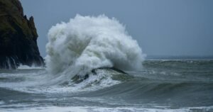 Dramatic scene of massive ocean waves crashing on a rocky coastline under a cloudy sky.