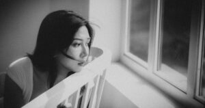 Black and white depressed ethnic female sitting on chair and looking out window at home