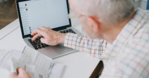 Elderly man at desk handling finances online with documents and laptop.