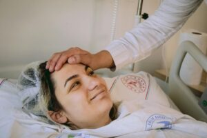 Major Surgery May Be Harming Memory in 1 in 7 Older Adults Smiling woman in hospital bed with supportive hand on forehead, conveying warmth and care.