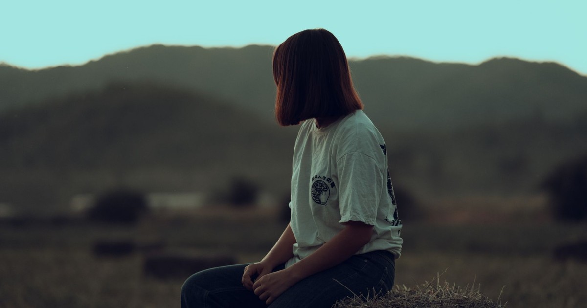 A woman in a white t-shirt sits on a hay bale, overlooking a tranquil, mountainous landscape.