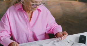 These 10 States Pay Retirees the Biggest Social Security Checks Elderly woman in pink blouse reading documents at a table indoors.