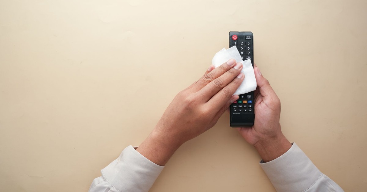 Hands cleaning a TV remote control with a cloth on a beige background for hygiene and cleanliness.