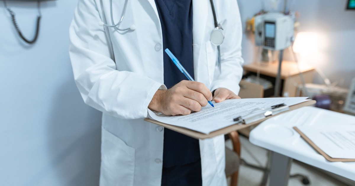 A doctor in a lab coat writing notes in a hospital setting, using a clipboard.