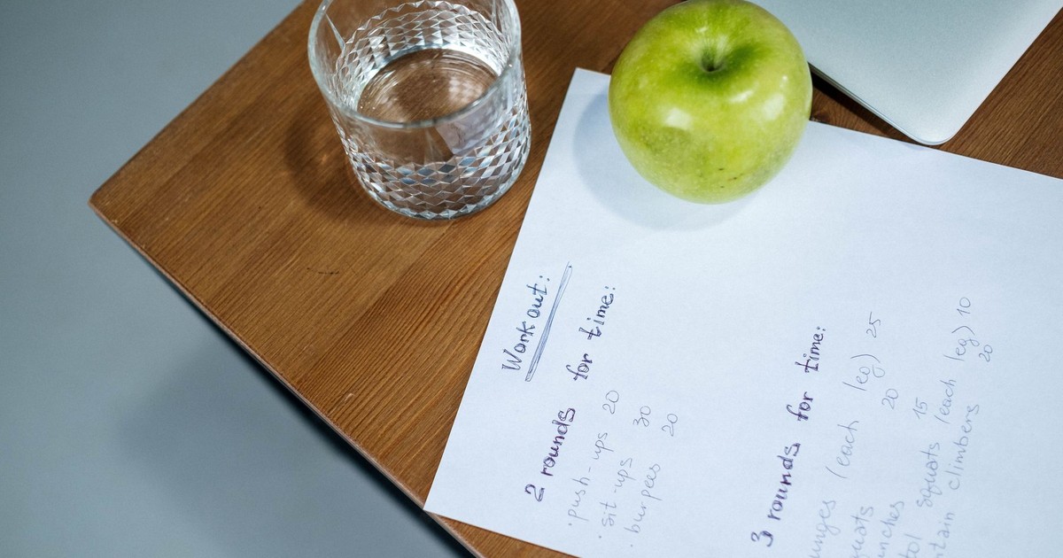 A green apple, glass of water, and workout plan on a wooden desk symbolize healthy living.