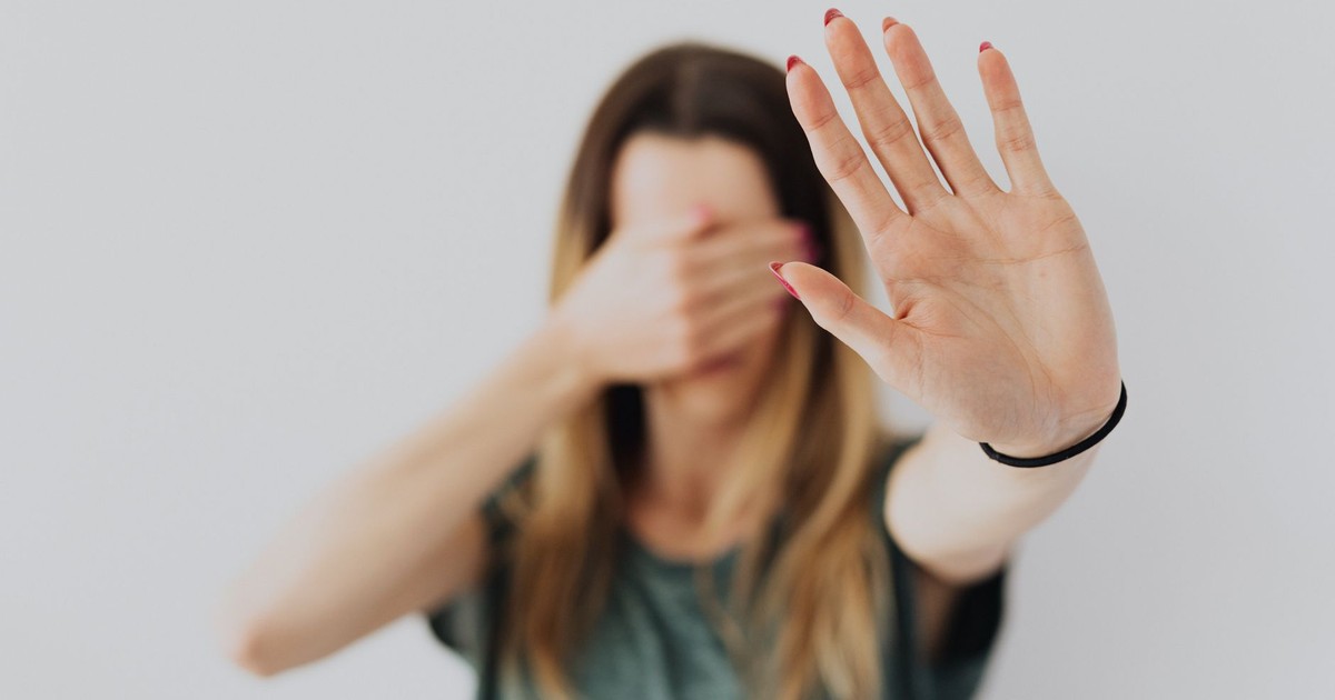 A woman in a gray shirt covers her face with her hand in a stop gesture, evoking a sense of fear and protection.