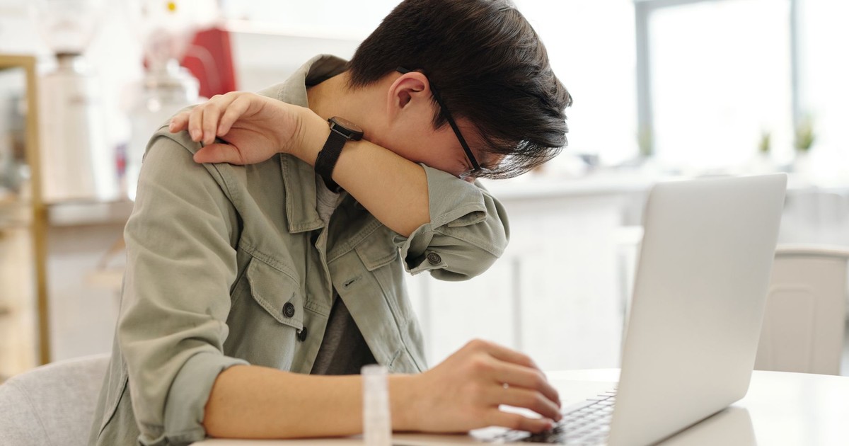 A man sneezes into his elbow while using a laptop, depicting common cold symptoms in a modern workspace.