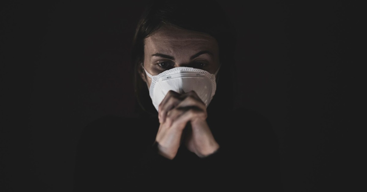 A woman wearing a protective face mask appears concerned in a dark setting, evoking pandemic safety themes.