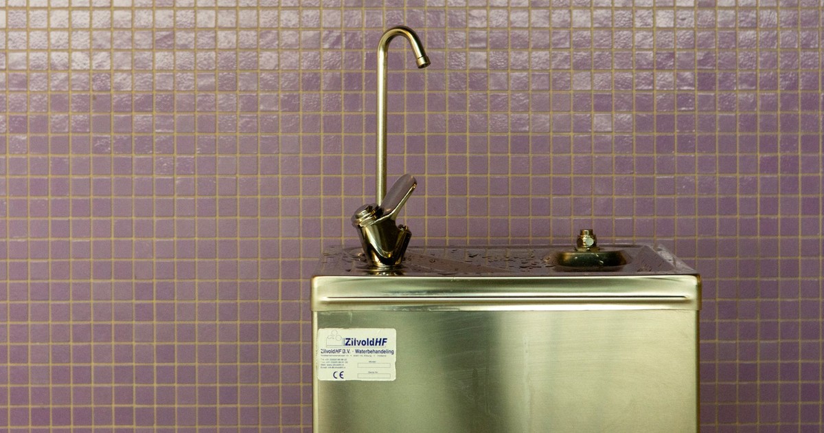A stainless steel drinking fountain set against a purple mosaic tiled wall, urban environment.
