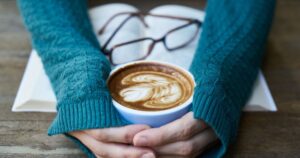 Drinking 5 Cups of Coffee Daily May Cut Your Liver Cancer Risk by Nearly Half Hands warming over a latte art coffee, with book and glasses, creating a cozy indoor ambiance.