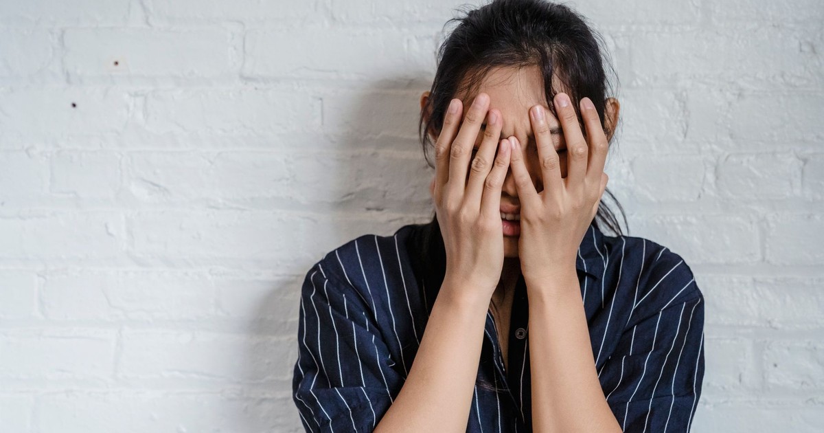 A young woman with hands covering face expresses stress against a white brick wall background.