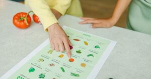 Not All Vegetables Are Created Equal: 9 Veggies That May Not Be Adding To Your Health Close-up of hands pointing to a vegetable nutrition chart with fresh tomatoes on the table.