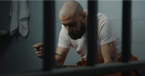 Inmate in tattoos in orange uniform sits on bed in jail cell, eats prison food from iron bowl. Male prisoner serves imprisonment term for crime in prison. Detention center or correctional facility.