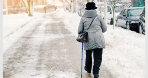 Elderly pedestrian using walking sticks for stability on slippery and hazardous snowy sidewalk in the city. Aged woman strolling on snowy sidewalk with walking sticks to avoid accidents, ensure safety