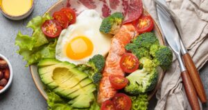 MIT Researchers Find Keto May Fuel Cancer in One Part of the Gut — But Prevent It in Another Balanced ketogenic diet meal with salmon, avocado, egg, bacon and vegetables on plate with cutlery top view on grey background