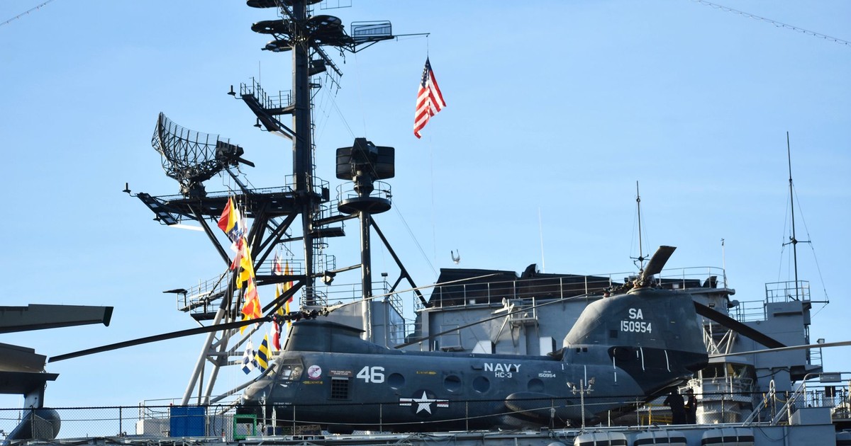 A US Navy helicopter is stationed on the deck of an aircraft carrier with radar equipment in the background.