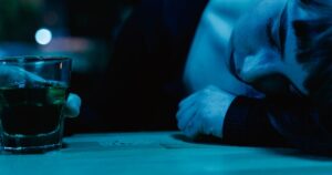 A woman rests on a table next to a glass of drink, illuminated in blue light.