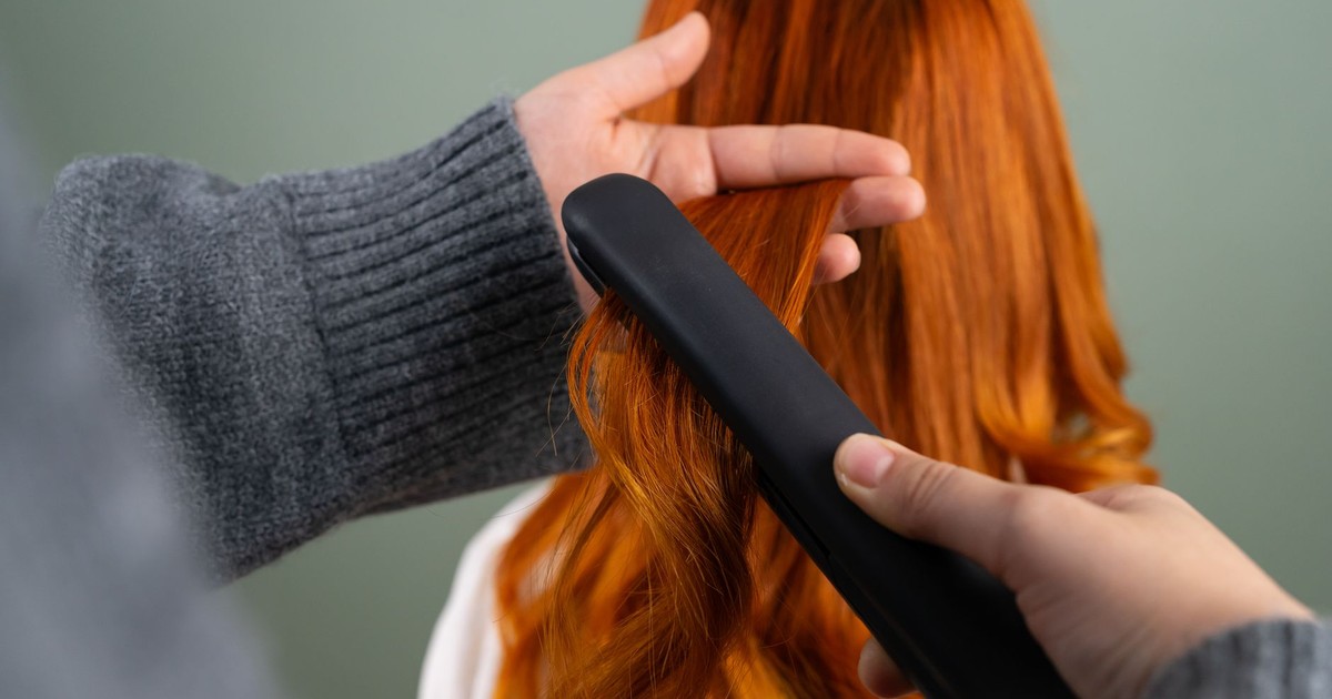 Hands styling long red hair using a flat iron indoors. Haircare and beauty concept.