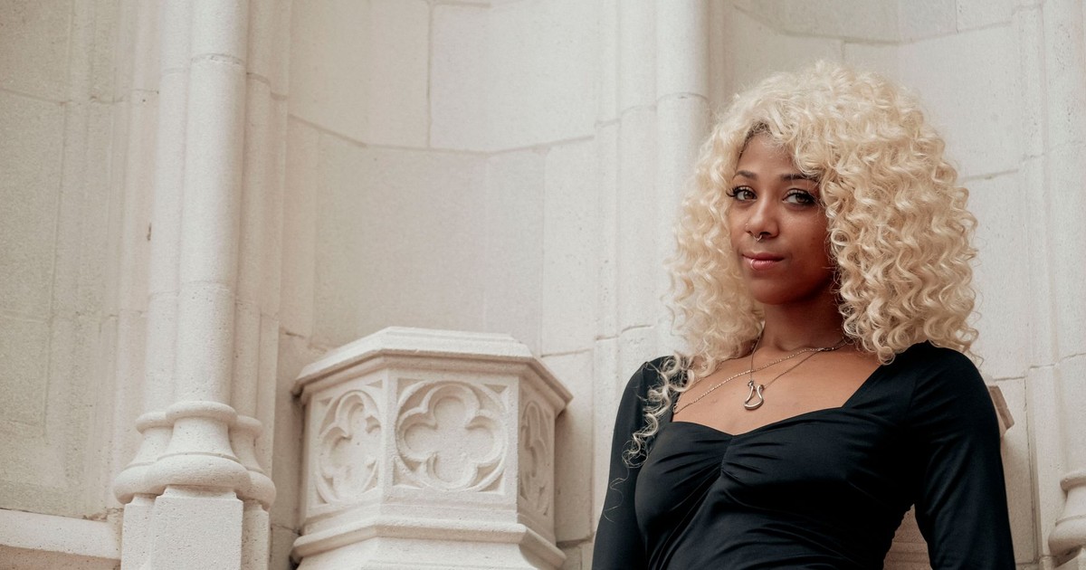 Elegant woman with blonde curls poses in front of detailed stone architecture, showcasing modern style.