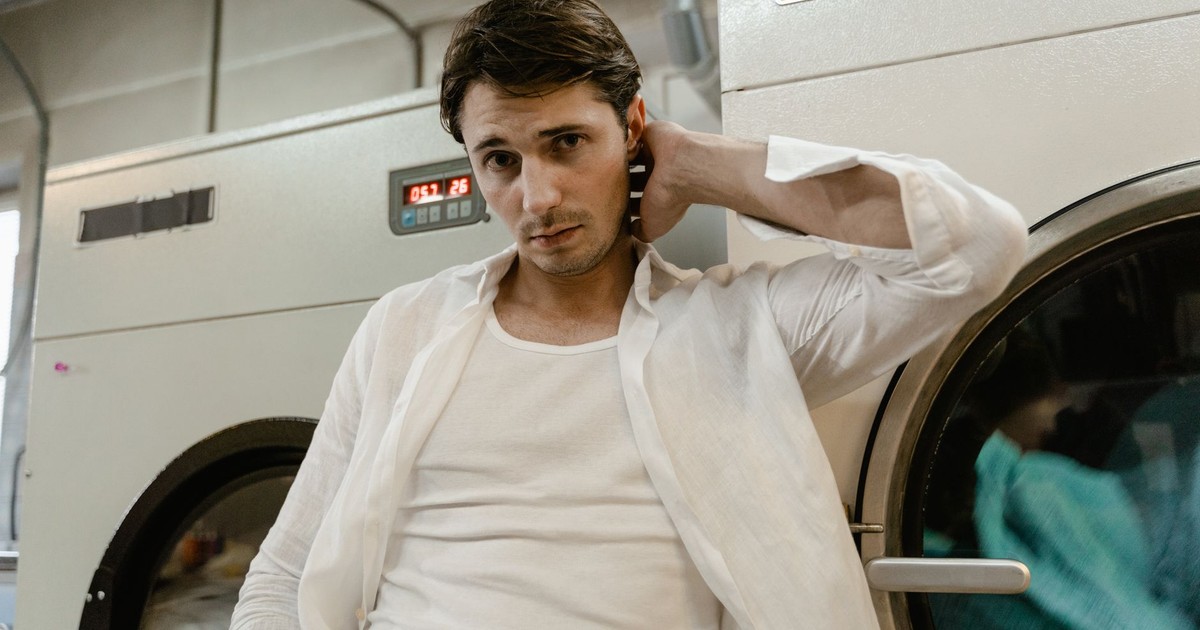 Young man in a white shirt leaning on a washing machine in a laundry facility.