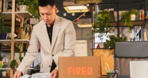 6 in 10 Companies Are Firing Gen Z Grads — Here's the Full Picture An employee packing documents into a box labeled 'FIRED' in an office environment.