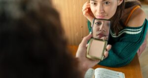 Girls Are Hitting Puberty Earlier Than Ever — Here's What's Behind It A young woman looks into a mirror while studying at a desk, reflecting thoughtfulness.