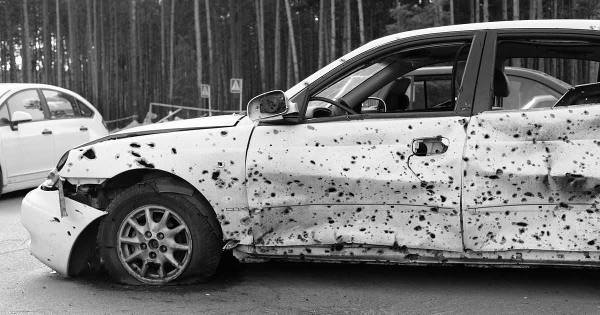 A monochrome image of a bullet-ridden car in a forest, conveying decay and abandonment.