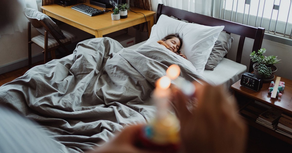 A father surprises his sleeping daughter with a birthday cake in a cozy bedroom setting.