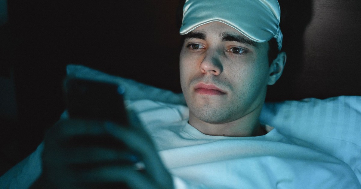 A man wearing a sleeping mask uses his smartphone while lying in bed at night.
