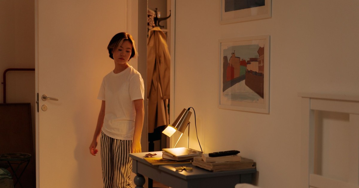 A person with short hair stands indoors next to a lit table in a cozy room.