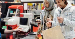 Science Backs the Man Who Always Skips Self-Checkout — Here's Why A Middle Eastern couple making a purchase at a supermarket checkout using a smartphone.