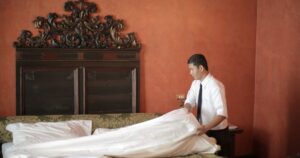 Should You Strip the Bed Before Checking Out? A Viral Debate Has Answers A hotel staff member in formal attire making a bed in a luxurious bedroom setting.
