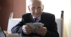Positive senior businessman in formal suit and eyeglasses counting money bills while sitting at wooden table with cup of beverage and near opened laptop
