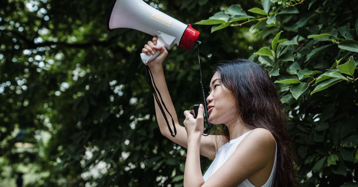 A young woman passionately speaks into a megaphone in a lush green outdoor setting.
